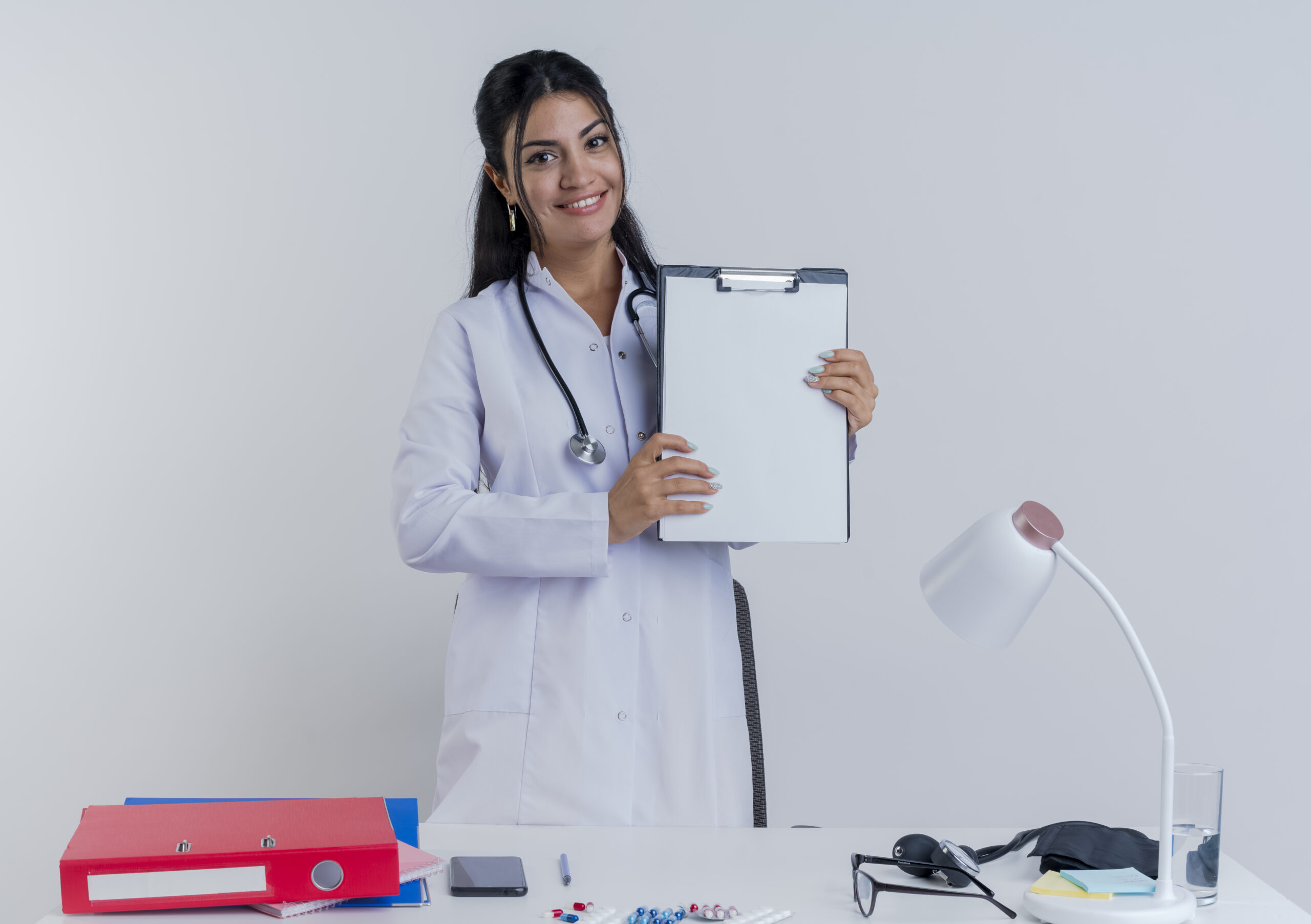 smiling young female doctor wearing medical robe and stethoscope sitting at desk with medical tools looking at camera showing clipboard to camera isolated on white background